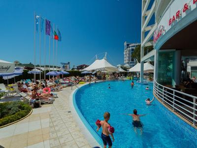 Piscina de hotel con bañistas, sombrillas y varias banderas bajo cielo despejado.