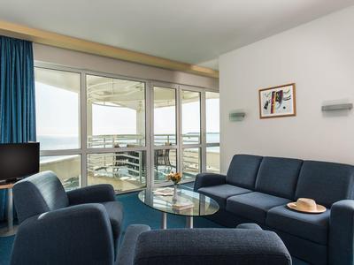 Modern hotelkamer met blauwe zitmeubelen, glazen tafel en grote ramen met uitzicht op de stad.