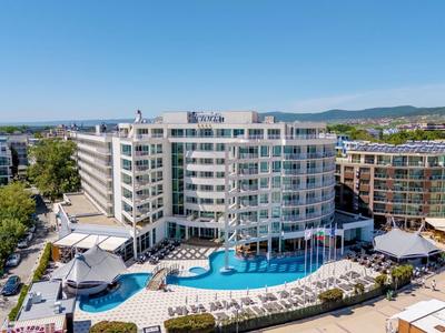 Een groot wit hotel met balkons rondom een blauw zwembad en ligstoelen onder een heldere lucht.