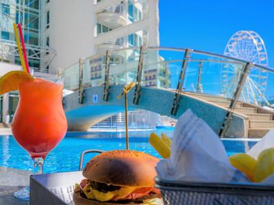 Hamburger en cocktail bij een zwembad met glazen brug en reuzenrad op de achtergrond.