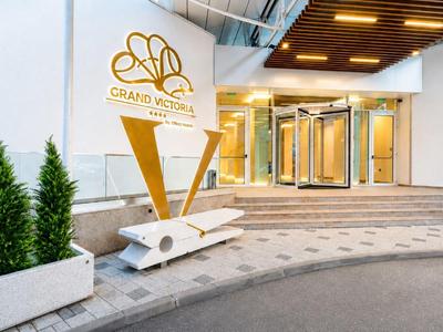 Wejście do hotelu z pozłacanym logo i nowoczesnymi szklanymi drzwiami, otoczone doniczkami z roślinami.