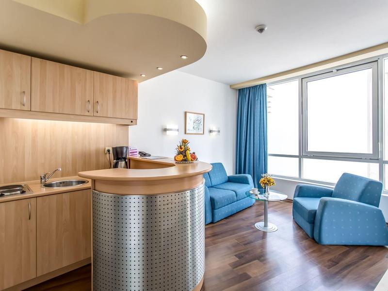 Moderne hotelkamer met blauwe zitmeubels, grote ramen en houten keukenhoek.