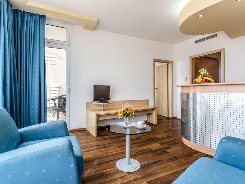 Lichte hotelkamer met blauwe fauteuils, houten vloer, glazen tafel en receptiebalie.