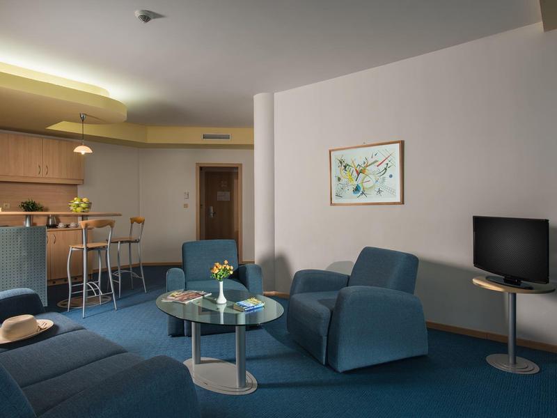 Hotelkamer met blauwe fauteuils, een bank, glazen tafels, een tv en een kleine keuken.