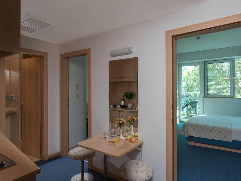 Compacte hotelkamer met kleine eethoek, keuken en slaapkamer met groot raam.