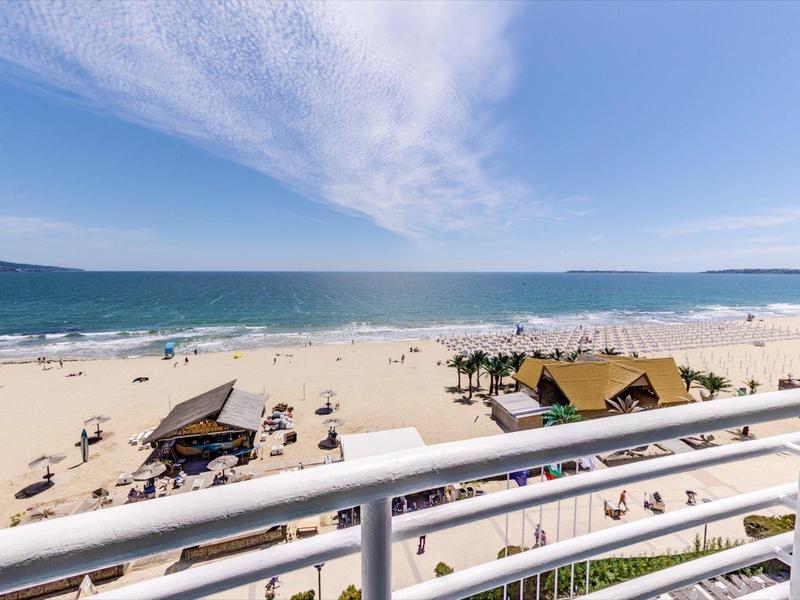 Zonnig strand met parasols, zonnebaders, ligbedden en rustige zee achter een wit hek.
