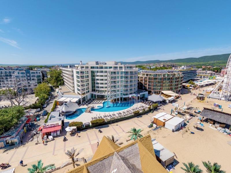 Kusthotelgebied met zwembad, strand, reuzenrad en zandzitplaatsen onder heldere lucht.