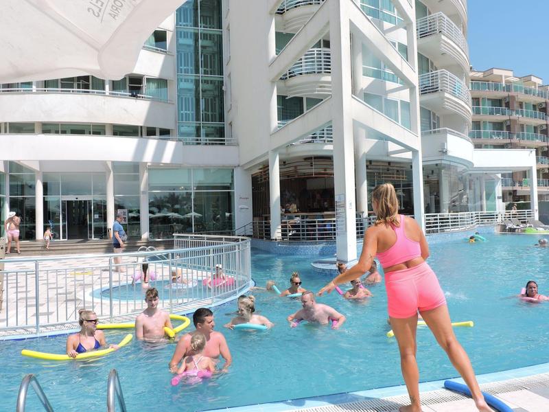 Buitenzwembad van hotel met gasten die zwemmen en instructeur die wateroefeningen geeft bij zonnig weer.