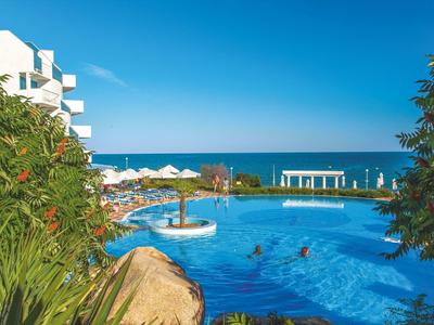 Hotel pool with people, white sun umbrellas, and sea view on a sunny day.