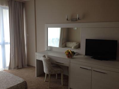 Bright hotel room with desk, armchair, large mirror, and TV next to window with curtains.