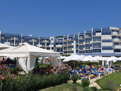 Modern multi-story hotel with garden and guests under sun umbrellas.