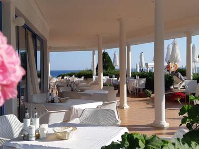Covered terrace with white tables and chairs, pink flowers, and sea view.