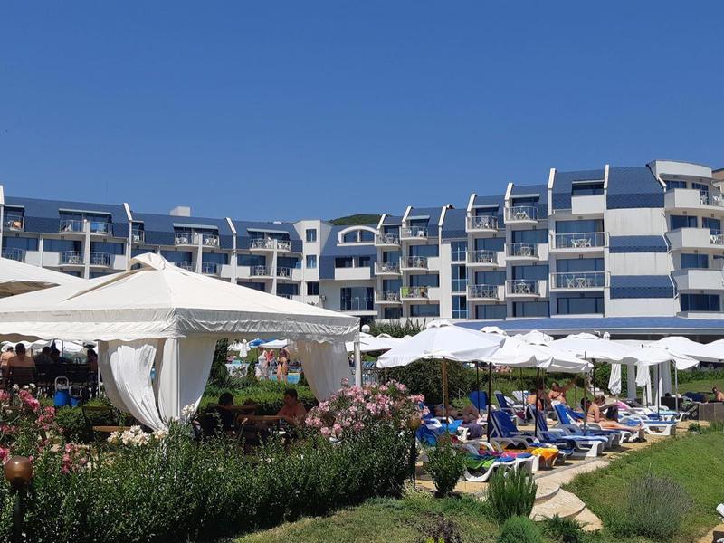 Modern multi-story hotel with garden and guests under sun umbrellas.