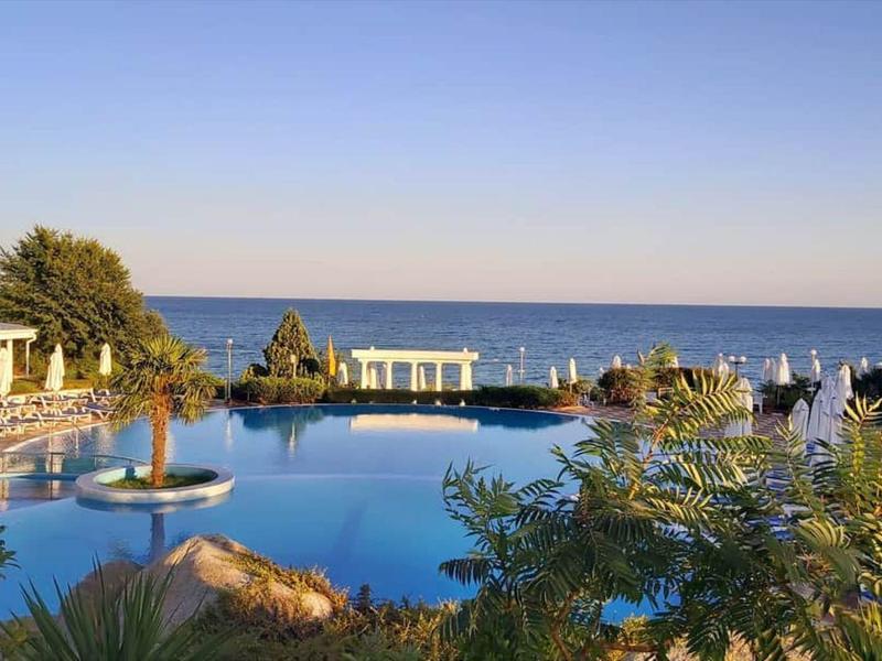 Large pool with water fountain and sun umbrellas overlooking the sea under a clear sky.