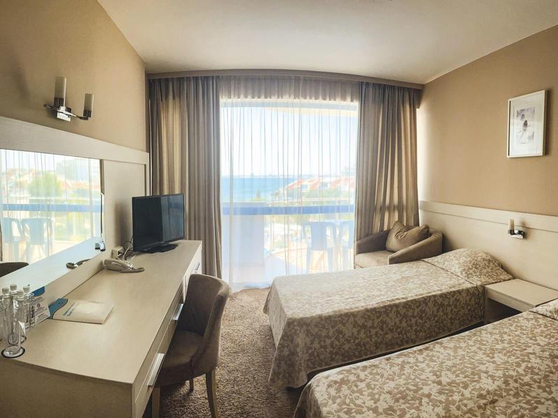 Hotel room with two twin beds, desk, chair, and water view through large window.