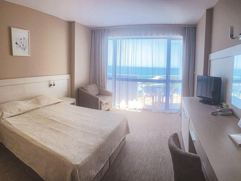Bright hotel room with double bed, desk, TV, and balcony with sea view.