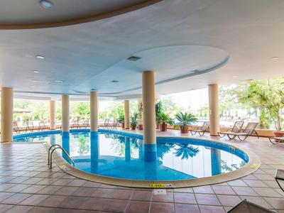 Covered hotel pool with columns, surrounded by lounge chairs and plants.