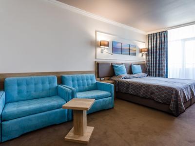 Modern hotel room with blue sofa, bed, carpeted floor, and large window with curtains.