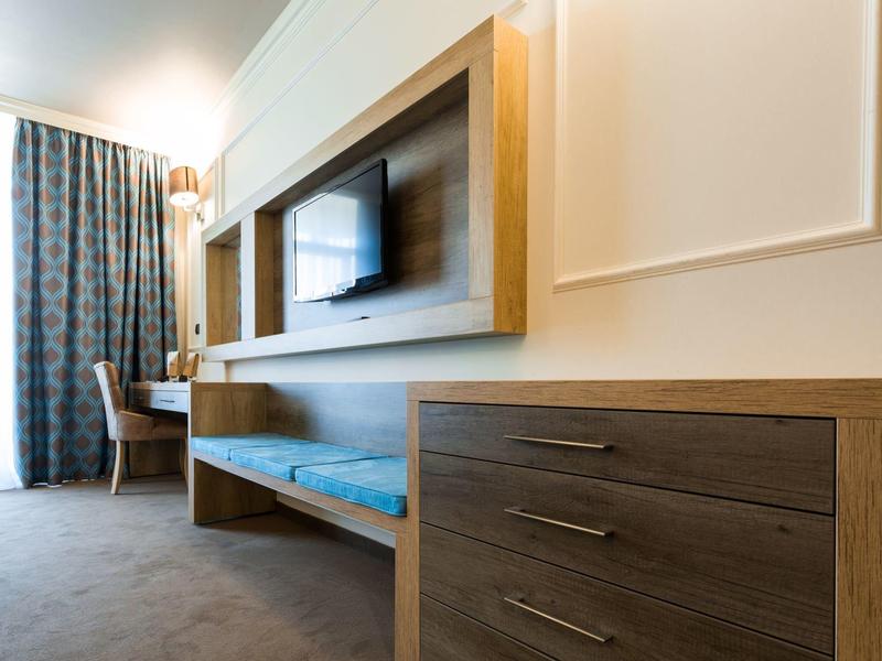 Modern hotel room area with wooden furniture, desk, and wall-mounted TV.