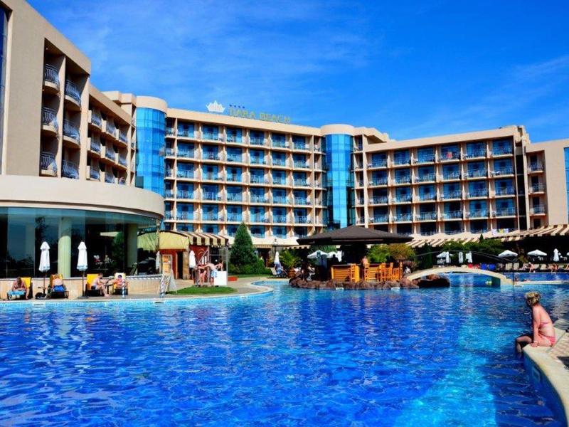 Modern hotel with large outdoor pool and sun terrace under clear blue sky.