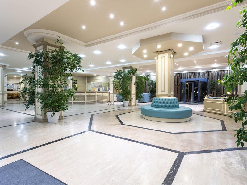 Bright hotel lobby area with seating, columns, and plants under built-in ceiling lights.