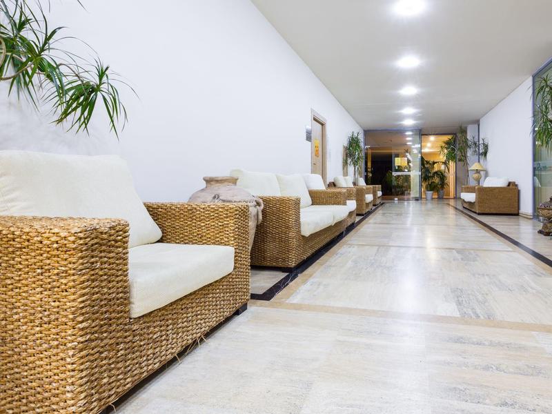Long hotel corridor with rattan armchairs and white cushions on the left side.