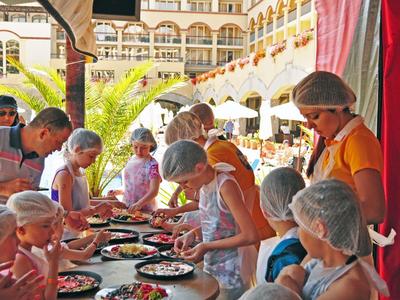 Kinderen met haarnetjes maken pizza's aan een zonnige buitentafel in een binnenplaats met bogen.