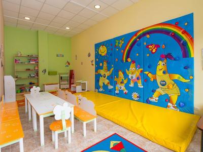 Lichte speelkamer voor kinderen met kleurrijke muurschildering, gele bank en witte tafels en stoelen.