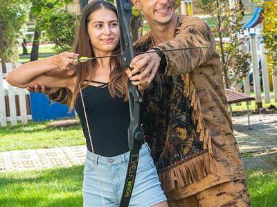 Een stel oefent boogschieten buiten op een zonnige dag, omgeven door groen gras en bomen.