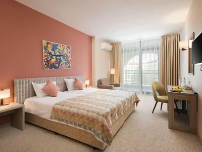 Helles Hotelzimmer mit Doppelbett, buntem Wandbild, Schreibtisch und großem Fenster mit Vorhängen.