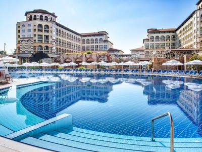 Großer Hotelpool mit Sonnenschirmen und mehrstöckigem Hotelgebäude im Hintergrund unter blauem Himmel.