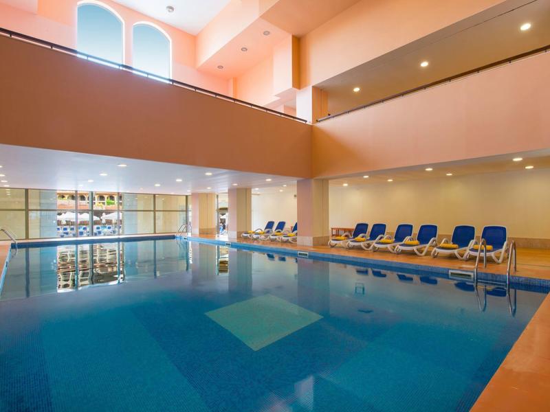 Overdekt zwembad met ligstoelen onder grote ramen in een ruime hotelruimte.