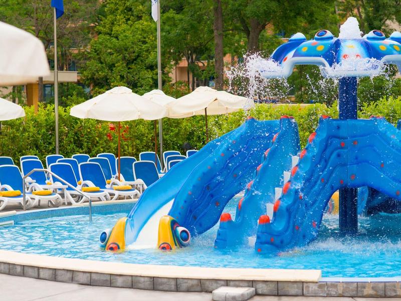 Kinderzwembad met blauwe schildpadglijbanen en parasols bij een zonnig hotel buitenterrein.