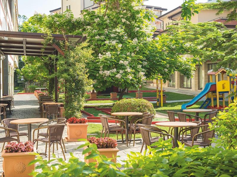 Buitenterras met tafels en stoelen naast een tuin en een kinder speelplaats.
