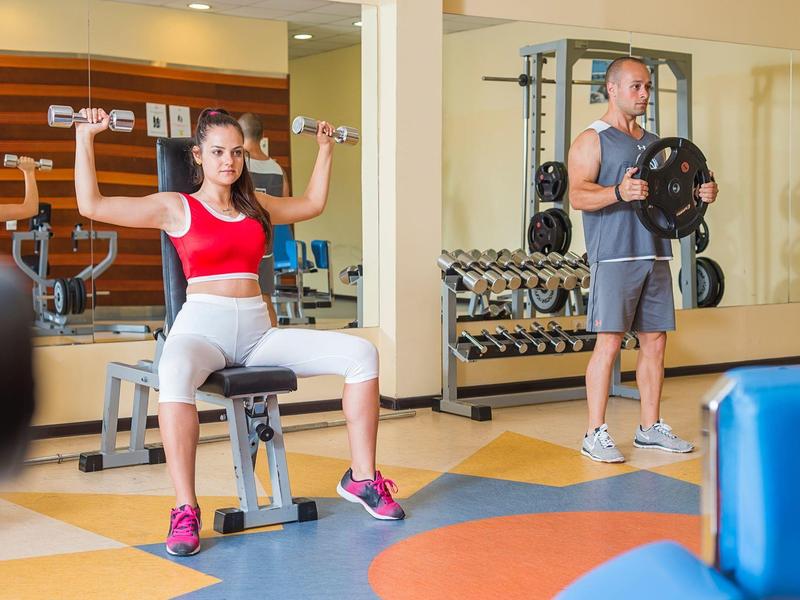 Man en vrouw trainen met gewichten in een sportschool met spiegels en halterrekken.