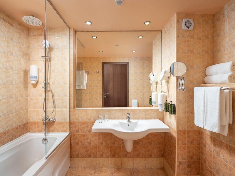 Hotelbadkamer met bad, wandgemonteerde wastafel, grote spiegel, handdoeken en beige tegels.