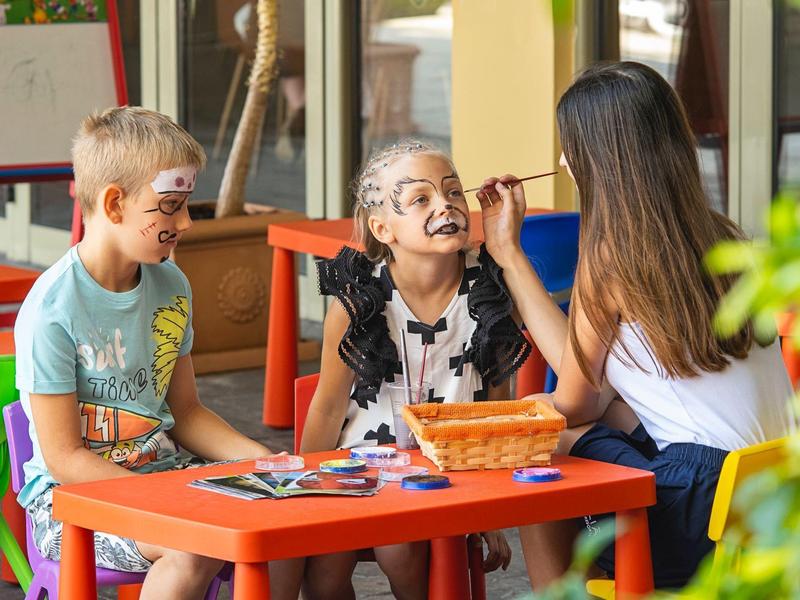 Twee kinderen laten zich schminken door een vrouw aan een klein buitentafeltje.