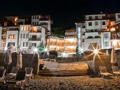 Nachtaufnahme eines beleuchteten Hotels mit Liegestühlen am Strand im Vordergrund.