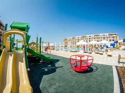 Children's playground with slide and merry-go-round next to pool and hotel buildings under clear blue sky.