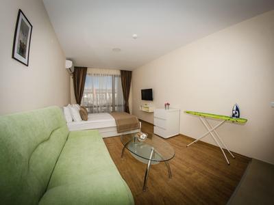 Bright hotel room with bed, green sofa, and ironing board on wooden floor.