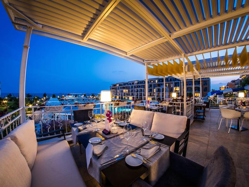 Evening rooftop terrace of a hotel with seating areas and views of illuminated buildings by the water.