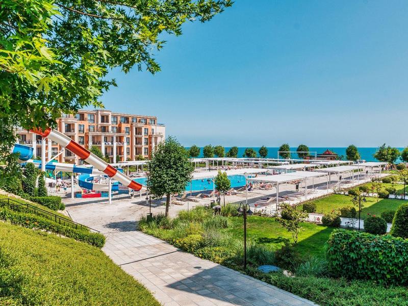 Large outdoor pool with slide and lounge chairs next to a hotel building and green gardens.