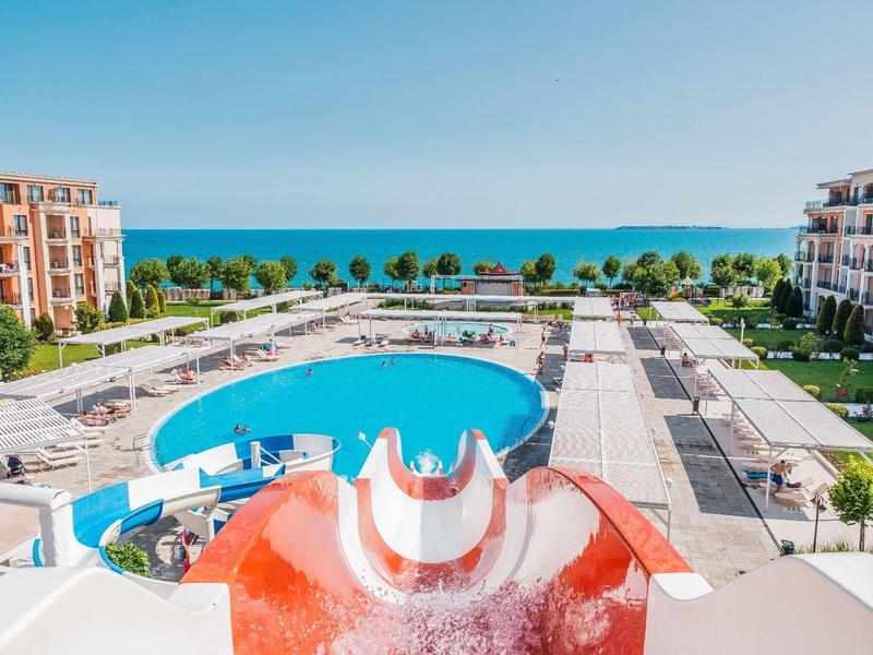View of holiday resort with pool, water slide, and sea in the background under clear sky.