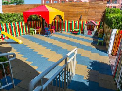Bunter Kinderspielplatz mit Rutsche, Pavillon, Zaun und farbigem Bodenmuster bei Sonnenschein.