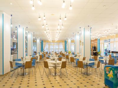 Großer heller Speisesaal mit vielen Tischen, Stühlen und hängenden Glühbirnen an der Decke.