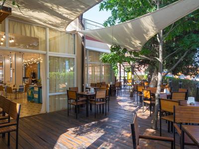 Außenbereich eines Cafés mit Holzboden, Tischen, Stühlen und Sonnensegeln im Schatten.
