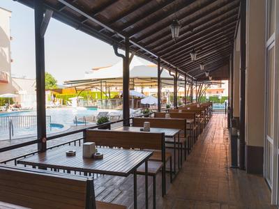 Überdachte Terrasse mit Holztischen und Stühlen neben einem Pool, sonnige Atmosphäre.
