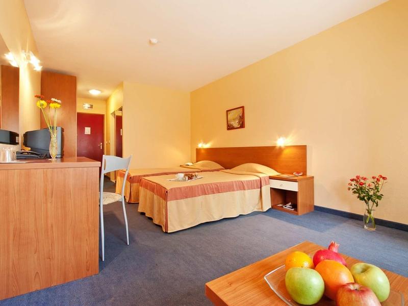 Bright hotel room with double bed, desk, chair, and fruit on a table.