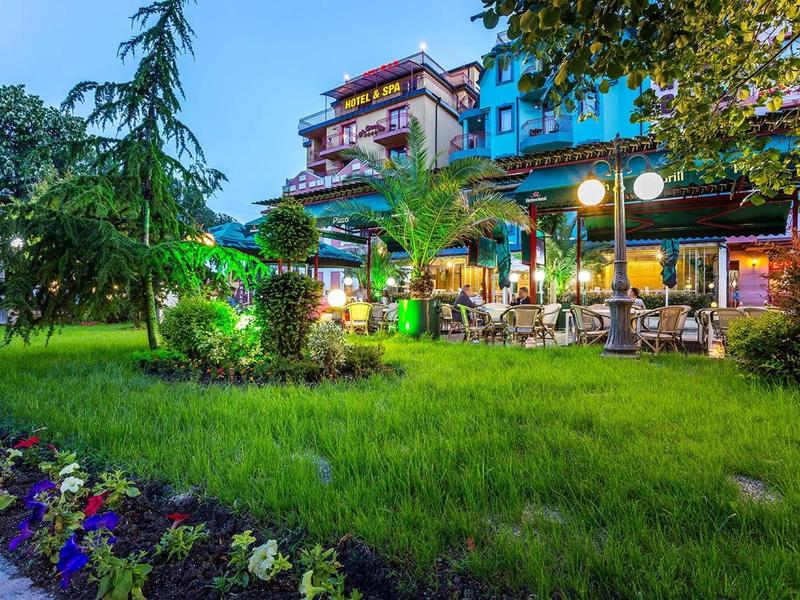 Illuminated hotel with terrace and green lawn with flowers at dusk.