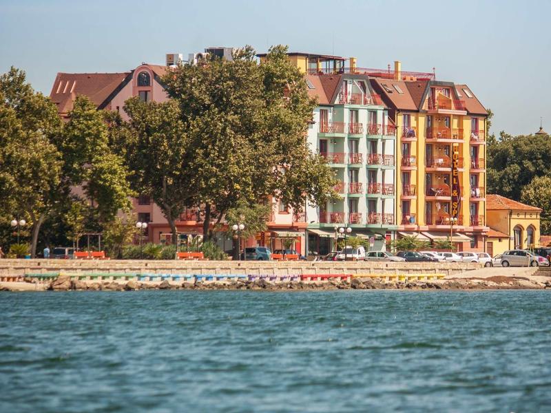Multi-story hotel building by the waterfront with sun loungers and trees in front.